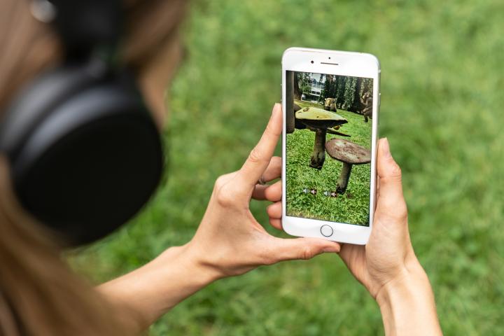 Zu sehen ist der Ausschnitt einer jungen Frau auf einer Wiese, die ein Handy in der Hand hat, auf dessen Screen Pilze auf der Wiese erscheinen.