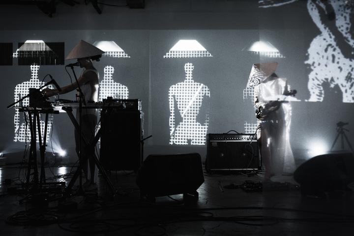 The Duo group A is performing live on stage, in the background there are visuals with abstract figures wearing the traditional asian cone-hat