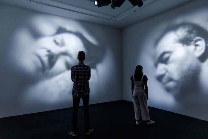 In a dark room, videos of two sleeping people are projected onto the wall. In front of them are two viewers.