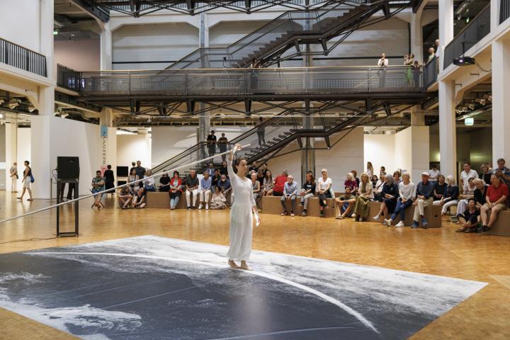 Performance of Ulrike Rosenbach's "Die einsame Spaziergängerin" (The Lonely Walker), the artist, dressed all in white, holds a pole and walks in a semicircle along a path while being filmed. In the background you can see a large audience.