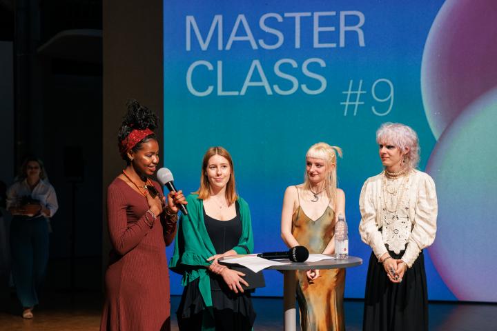 Four people on stage at the [MASTERCLASS] #9 opening. The person on the far left is holding a microphone in one hand and has the other hand on her heart.