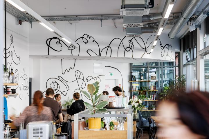 In the picture you can see the artwork of Alina Bukina "One Line Series", in intro Café at Kronenplatz. A black line runs through the entire cafe on the walls. In the cafe itself sit some people.