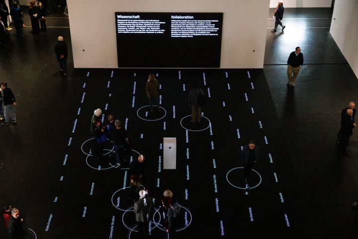 You can see several people on a black surface. On this surface there are words. Around the people are white circles. In the background are two screens.
