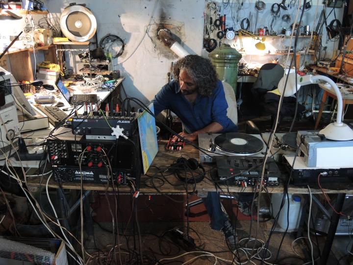 Zu sehen ist ein Mann in einem Raum voll Kabel und Geräte, der Musik produziert. 