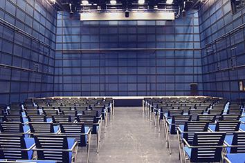 Bild des ZKM Medientheaters mit Reihenbestuhlung und Bühne. Moderne Architektur in blauem Grundton.