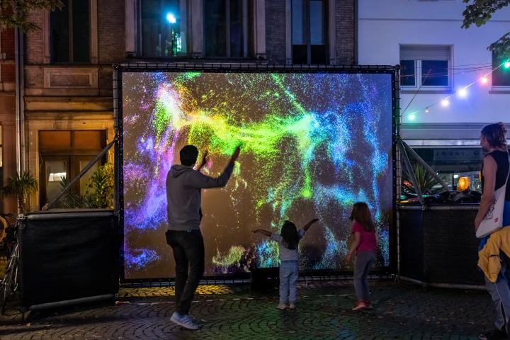 In der Fußgängerzone steht eine Leinwand mit leuchtend bunten Farbpartikeln, davor bewegen sich Erwachsene und Kinder.