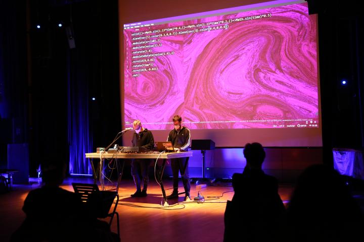 Das Foto zeigt zwei Männer die vor einer großen Leinwand stehen und ein Pult mit Laptop vor sich aufgebaut haben. Auf der leinwand sieht man groß einen rosanen Hintergrund und weiße Schrift. Ein paar Leute aus dem Publikum sind von hinten zu sehen.