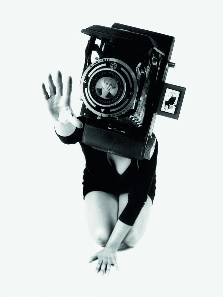 The black and white photo shows a woman in a black mini dress on her knees. Her face is covered by an oversized camera. 