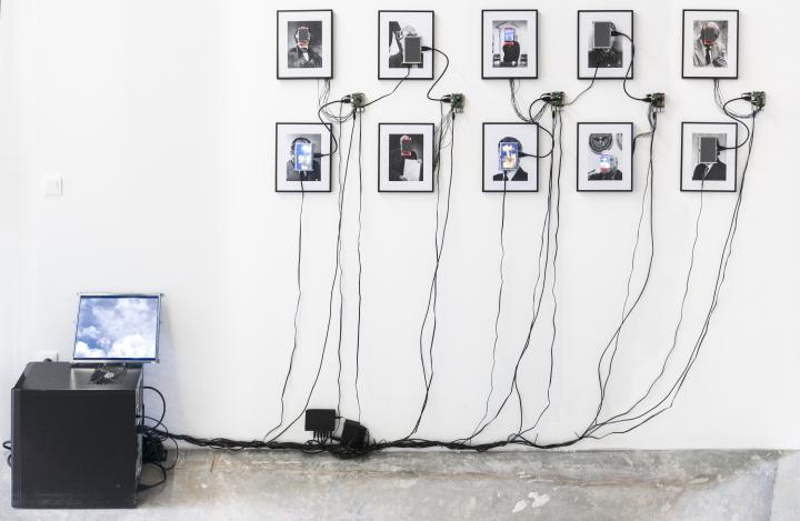 The photo shows 10 framed portraits with small monitors on the shown faces. These monitors are connected to a computer by cables.