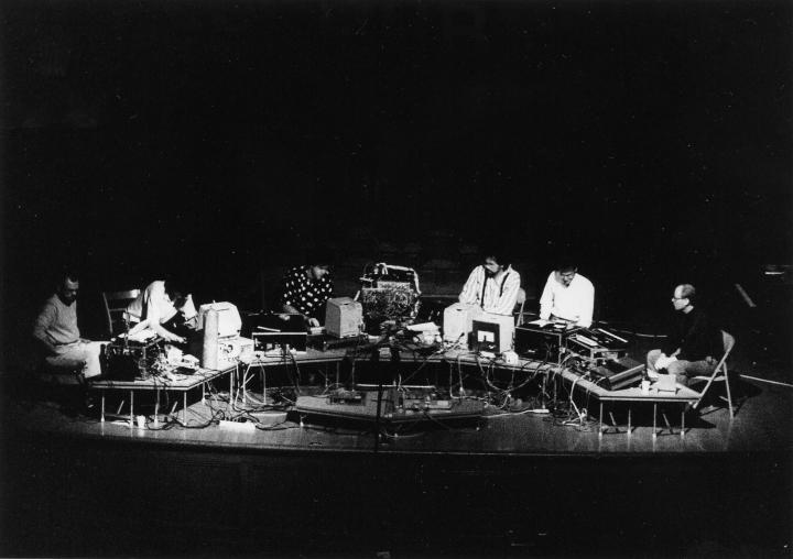 A black and white photo of the musician group »The Hub« from 1989, they are sitting in front of a long table with tube screens and many cables.