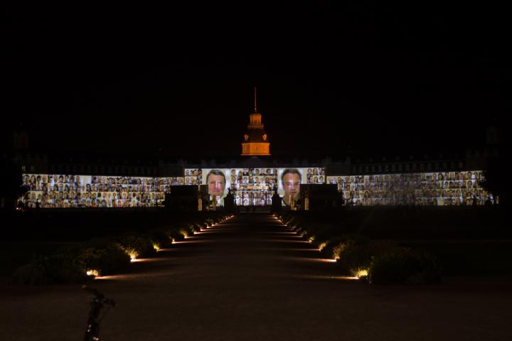  The facade of Karlsruhe´s Castle is projected with many passport photographs of various people.