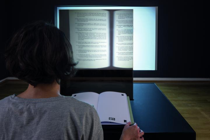You can see the back of a person's head, and in front of it is an open book. On the wall opposite is a projection of an opened book.