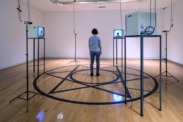 A room can be seen in which four monitors stand on a rack. A circle is glued to the floor, in the middle of which is a star and another circle. In the middle is a woman with her back to the viewer.