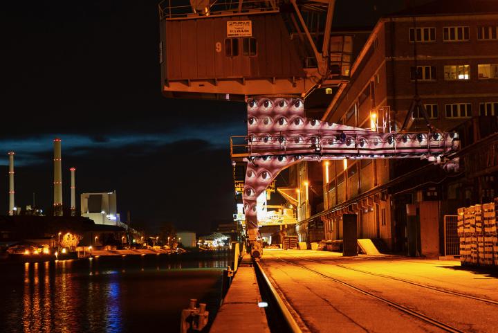 A port at night. Many pairs of eyes are projected on a column designed like an archway. The eyes look open in different directions.