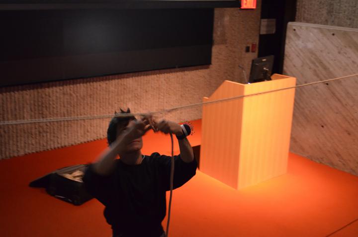 The photo shows a performer in the foreground who is hanging from a rope and harassing another one. She is wearing a pincushion around her left wrist. The person dressed in black stands diagonally in front of the lectern on the orange carpet.