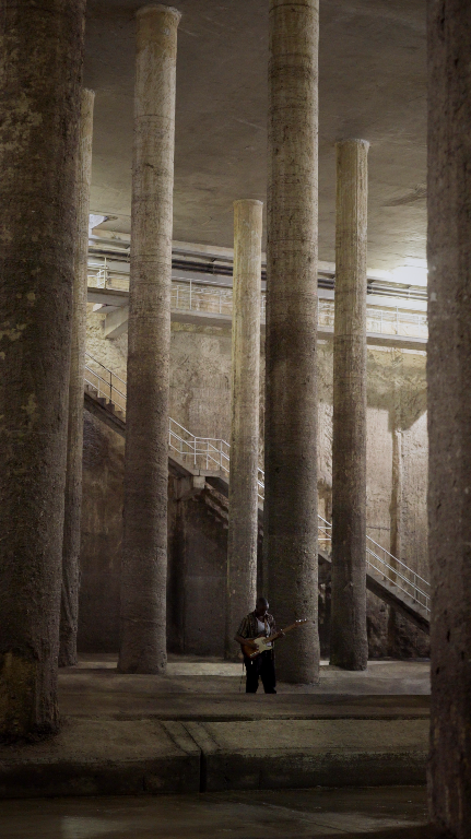 You can see a large dark columned room with a single guitarist standing and playing his guitar.