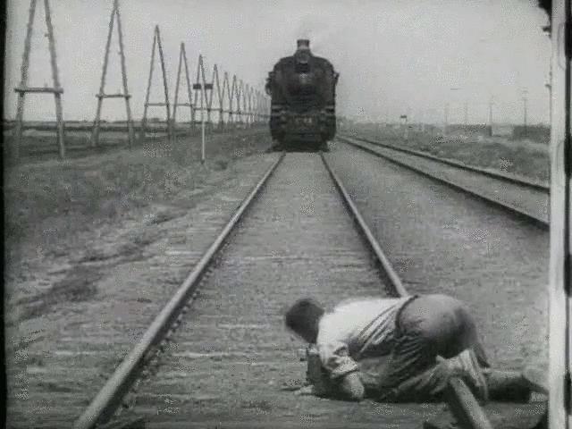The black and white screenshot shows a track with a railway. A man lies crouching over a track and looks in the direction of the upcoming train.
