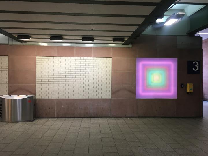 Zu sehen ist der Durchgang unter dem Karlsruher Hauptbahnhof. An der Wand ist ein leuchtendes Quadrat angebracht, das von innen nach außen einen Farbverlauf hat, der eine Tiefe erzeugt.