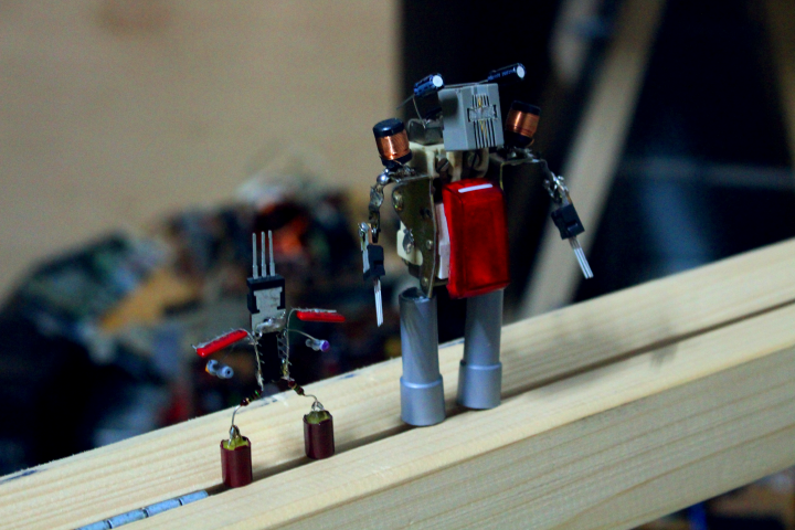Auf einem Holzbalken stehen zwei aus Elektronikschrott zusammengelötete Roboter.