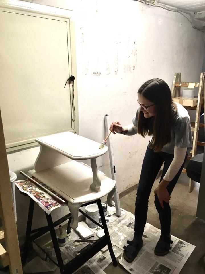 The picture shows a student using a brush to paint a wooden object with white paint in an interior room. The picture was created as part of the Cultural Academy Baden-Württemberg 2020/21.