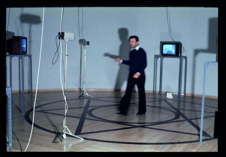 Ein Mann steht in einem Pentagramm umgeben von Monitoren und Kameras. Das Foto zeigt Peter Weibel im Rahmen der Ausstellung »respektive Peter Weibel«.