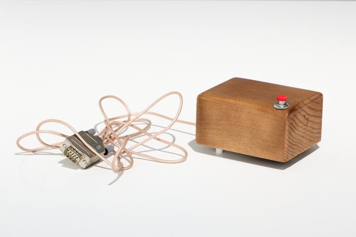 »Mouse« von Douglas C. Engelbart. Zu sehen ist ein Holzblock. Vorne am äußeren Rand befindet sich ein roter Knopf. An dem Holz ist ein Kabel mit Stecker befestigt.