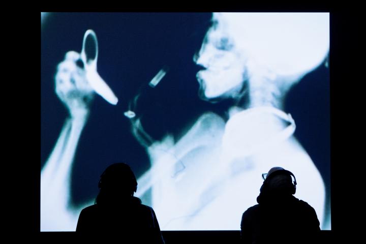 Man sieht das Werk »Sanctus«. Auf einem großen Bildschirm wird das Röntgenbild einer Person gezeigt. Davor stehen zwei Personen. 