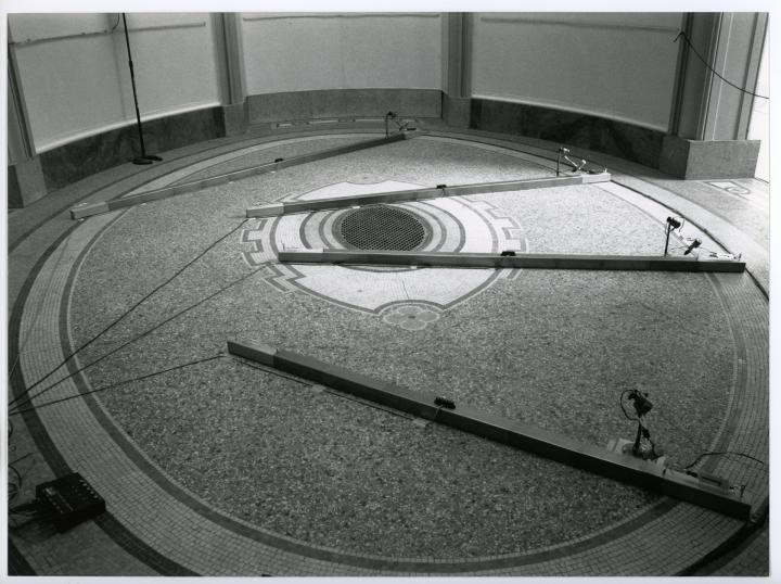 You can see the work »Number Made Audible«. The black and white photo shows a round room. On the floor are four long stilts to which cables are attached.