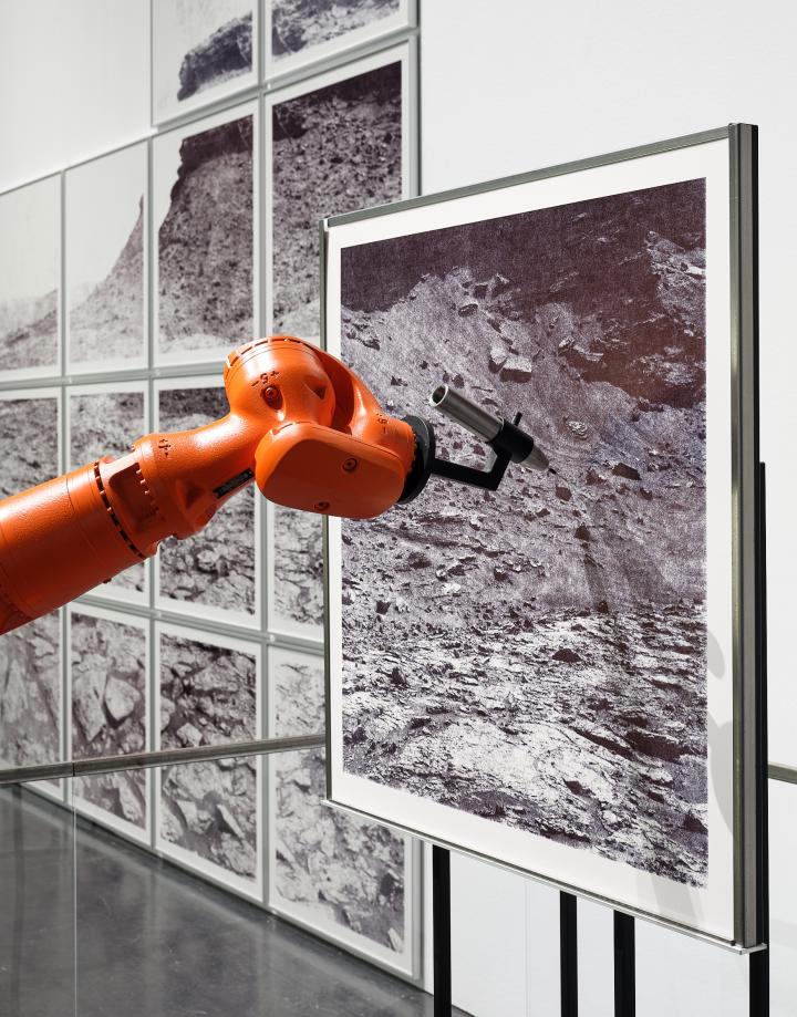»the native picture« von robotlab. Zu sehen ist ein orangener Roboterarm, der ein schwarz-weiß Bild von einer dürren Landschaft zeichnet.