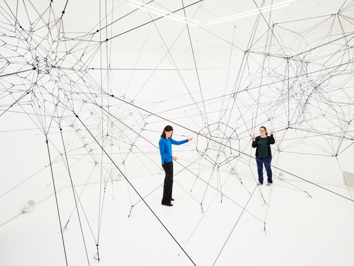 »Algo-r(h)i(y)thms« von Tomás Saraceno. Zu sehen ist ein großes Netz aus Fäden in einem weißen Raum. Zwei Personen stehen in diesem Raum und berühren diese Fäden.