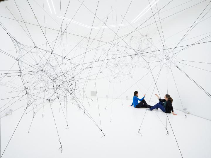 »Algo-r(h)i(y)thms« von Tomás Saraceno. Zu sehen ist ein großes Netz aus Fäden in einem weißen Raum. Zwei Personen sitzen in diesem Raum und berühren diese Fäden.