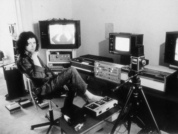 Das Foto ist schwarz-weiß und zeigt Ulrike Rosenbach, die Medienkünstlerin, in ihrem Studio in den 70er Jahren. Ulrike Rosenbach sitzt auf einem Drehstuhl und ist umgeben von Kameraequipment und Röhrenbildschirmen. 