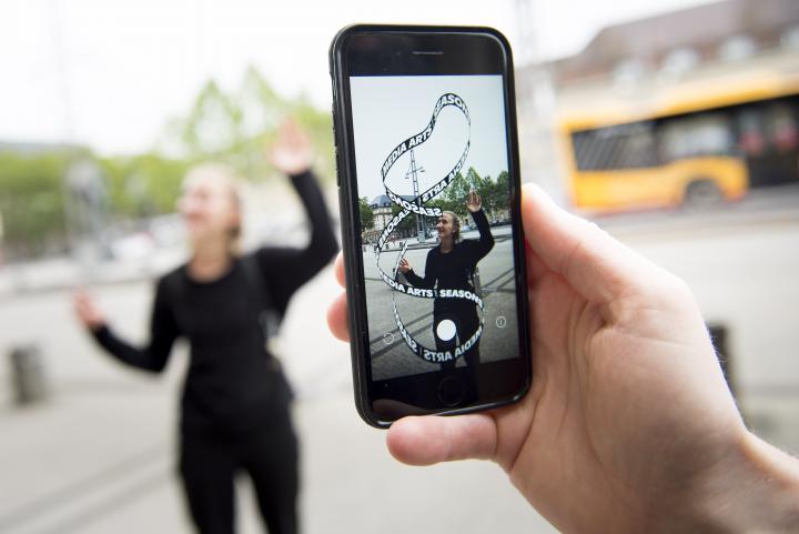 In the foreground you can see a mobile phone with an app artwork on its display. The virtual "Seasons of Media Arts" loop and a blonde woman trying to hold the ends of the loop together.