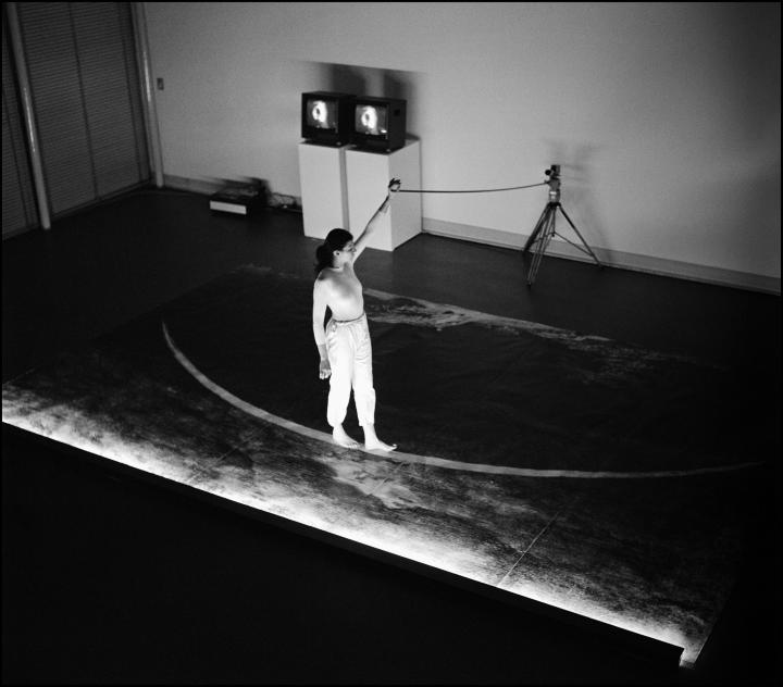 Ulrike Rosenbach, »The Lonely Walker«, 1979.You can see a person on a stage walking barefoot on a curved line. She is holding a rope in her hand, which is attached to a tripod. The image is black and white. 