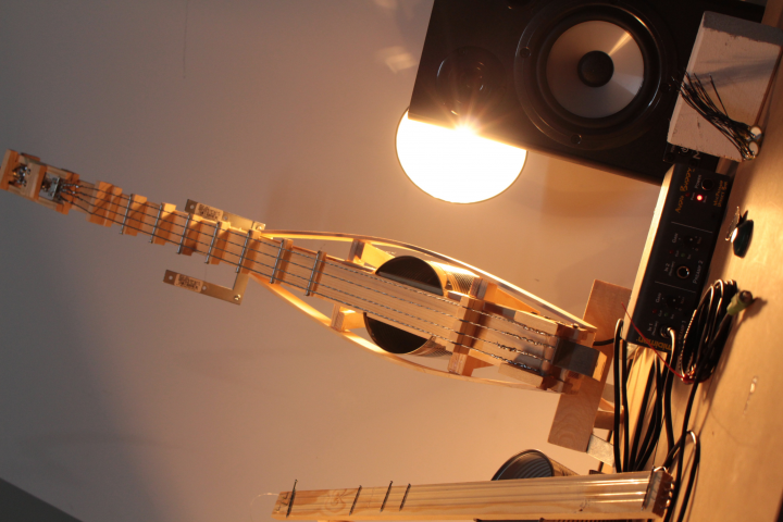 A diy guitar out of thin wood and a tin can is standing upright next to a speaker. A bright light is shining on the instrument from behind.