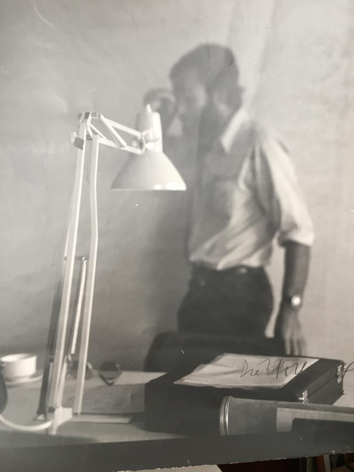 A black and white photograph shows sharply a desk lamp in the foreground. In the background one is out of focus a man, Hansjörg Mayer with a beard and a white shirt.
