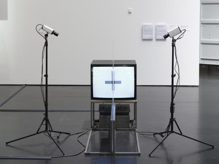 Das Foto zeigt einen Röhrenbildschirm der von zwei Kameras umrahmt wird. Die Kameras sind auf zwei Ständern montiert. Mittig zum Bildschirm ist auf einem Metallgestell eine Plexiglas Platte aufgestellt. Ein Kreuz leuchtet aus dem Rohrenbildschirm.  