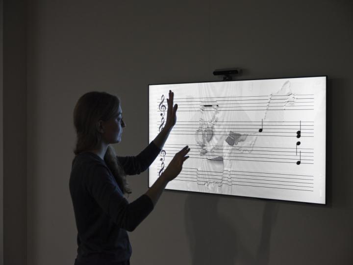 Das Foto zeigt eine Besucherin die mit ihren Händen über eine Noten Patitur fährt. Diese Notenpartitur wird von einem Bildschirm ausgestrahlt und simuliert.