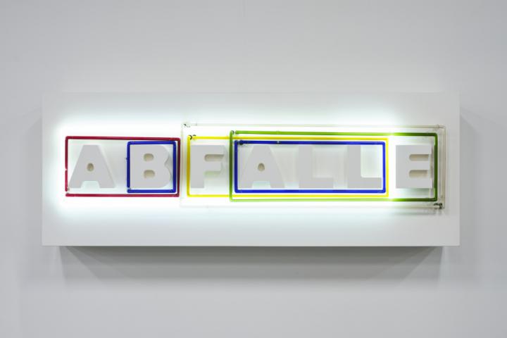 The photo shows white letters that form the word "WASTES". Around combinations of letters, neon tubes in different colors and sizes can be seen. These change their light conditions.