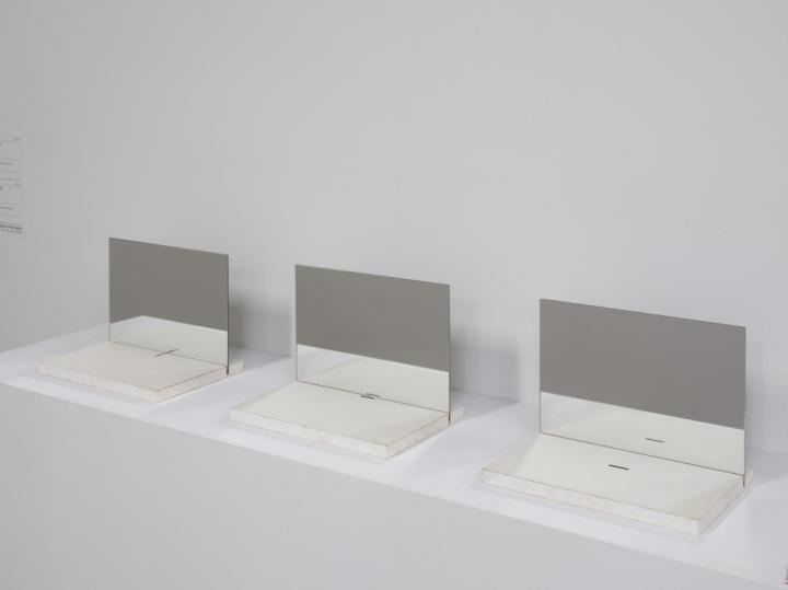 Das Foto zeigt eine dreiteilige Arbeit. Es liegen drei Papiere mit einer jeweils unterschiedlich angeordneten Linie auf Novopanplatten aufgelegt. Vertikal stehen Spiegel auf den Platten und spiegeln die dünne Linie auf dem Papier. 