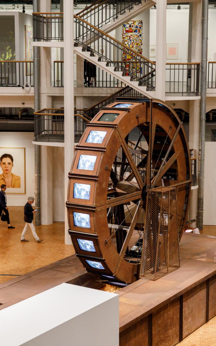 A huge metal mill wheel stands in an exhibition room. At the mill wheel there are tube screens on which videos of water are played.