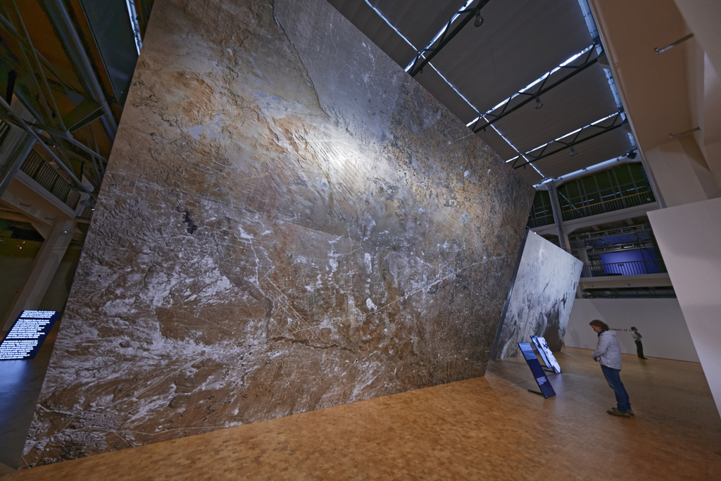 A person standing in front of a huge canvas on which a total of a cracked ground can be seen