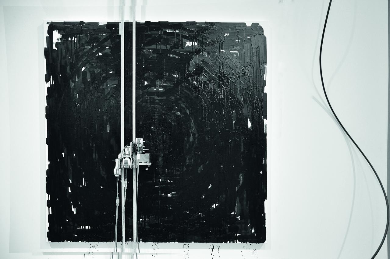 Eine Maschine malt schwarze Striche auf einer quadratischen weissen Fläche an der Wand