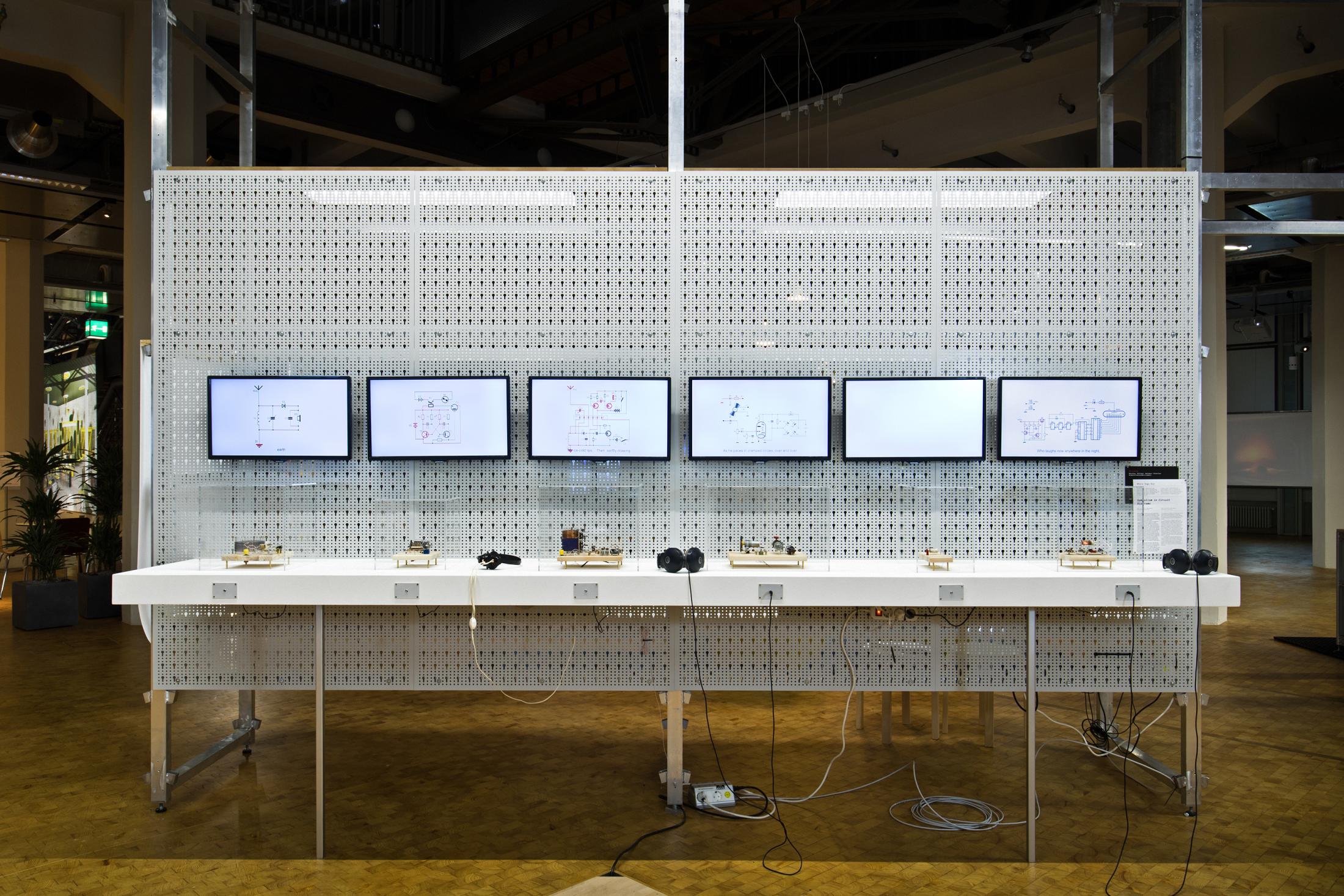 Das Foto zeigt sechs Bildschirme an einer weißen Gitterstruktur mit kleinen Objekten darunter und zwei Kopfhörern