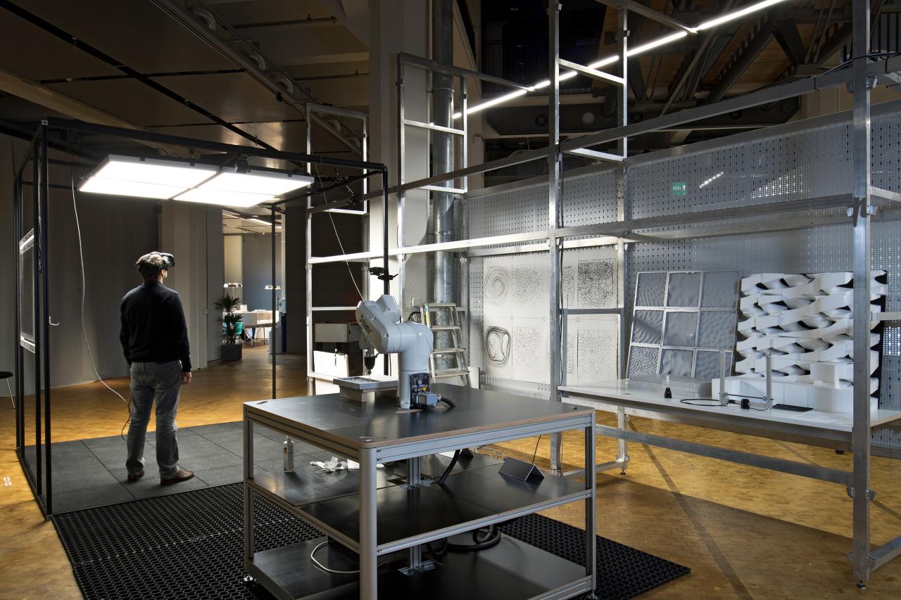 Installation with a robot and milled elements in grey shelves, next to which stands a man with VR glasses