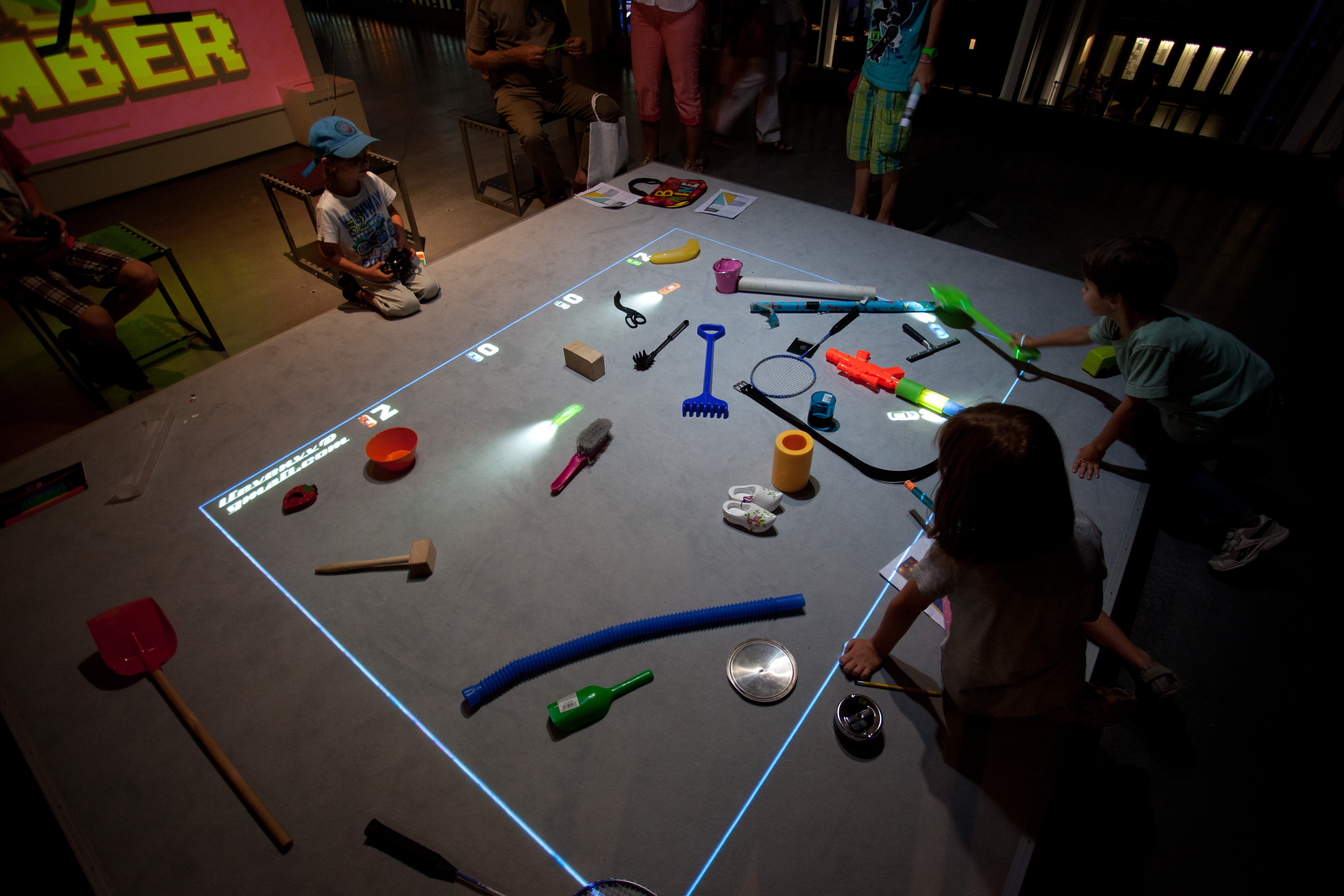Kinder sitzen vor einer Spielfläche auf der Gegenstände liegen und virtuelle Autos fahren