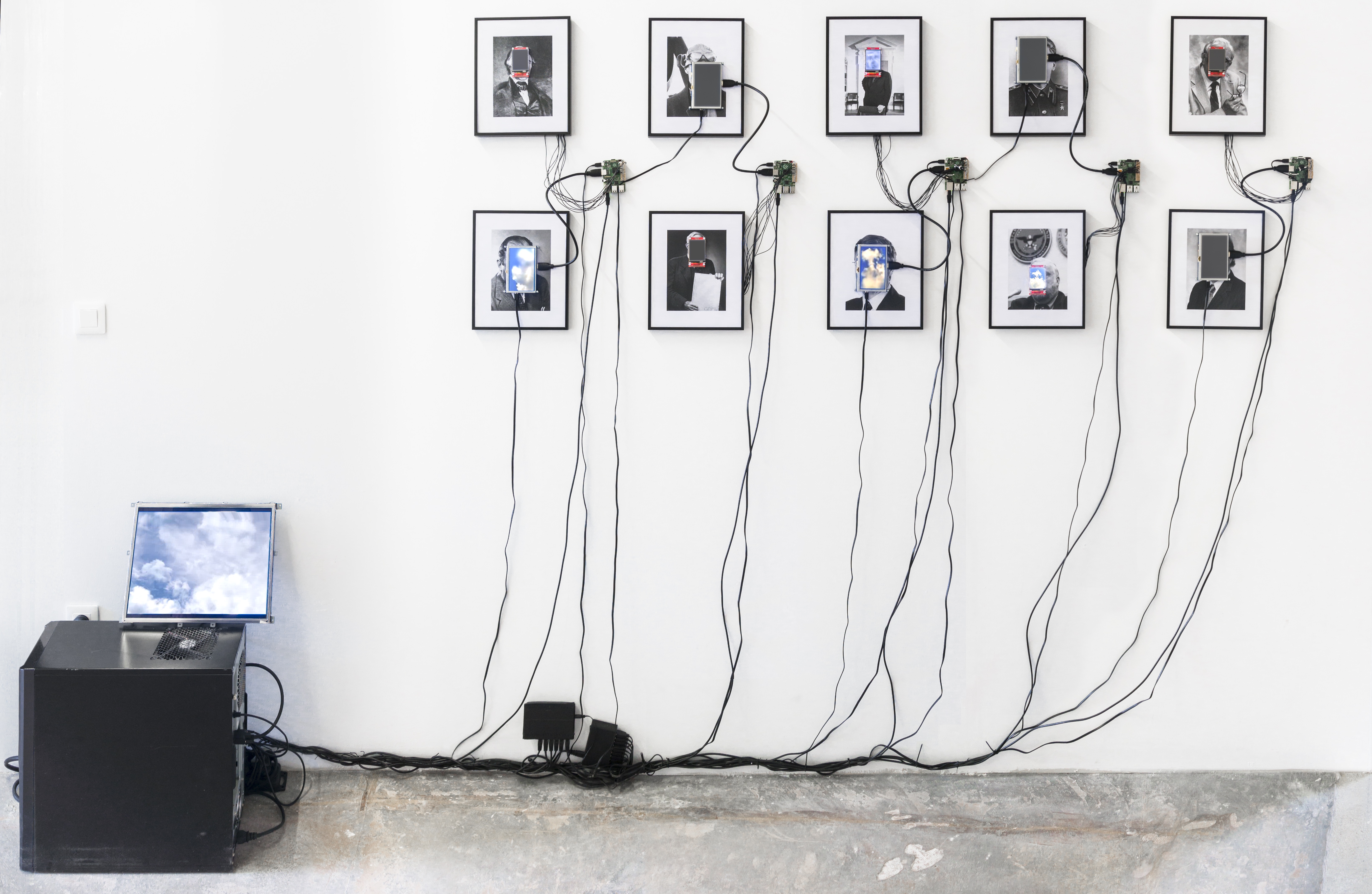 Das Foto zeigt 10 eingerahmte Portraits auf deren Gesichter kleine Monitore angebracht sind. Diese sind durch Kabel mit einem Computer verbunden.