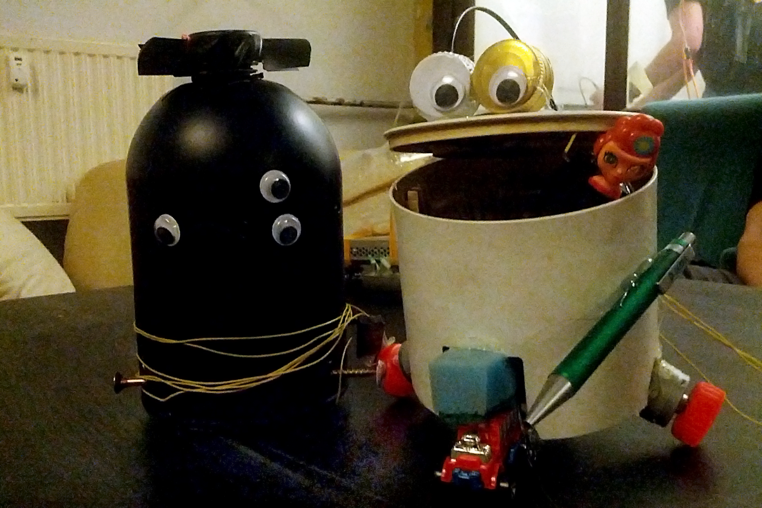 Two little robots that are made out of junk are sitting next to each other, one is black and has 3 eyes and one is made out of a bin with 2 eyes out of bottle caps, that are placed on the open mouth resp. lid of the bucket.