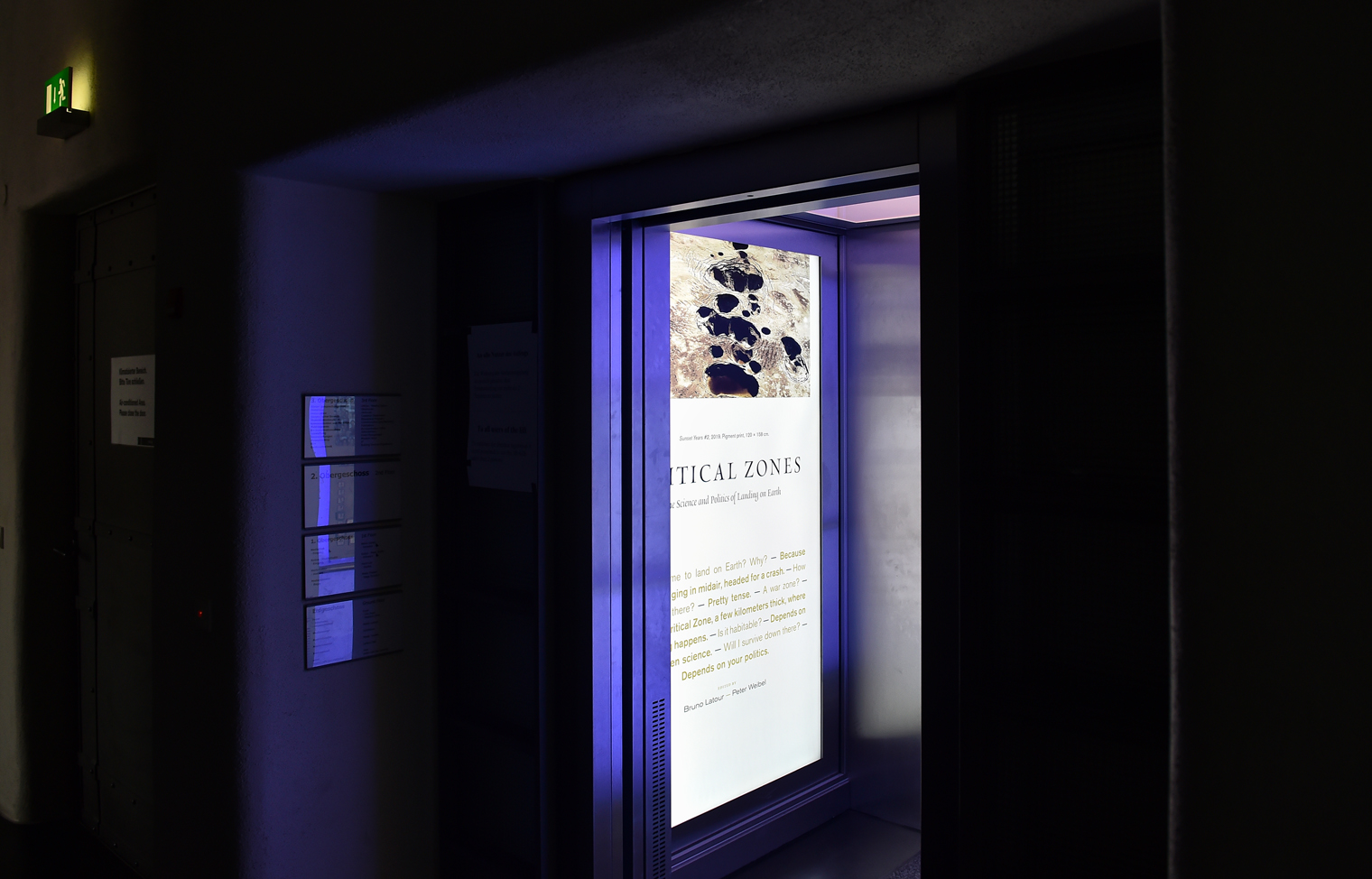 Zu sehen ist ein geöffneter Fahrstuhl. An der Innenwand des Fahrstuhls befindet sich ein großer Display, welcher einen Text zu der Ausstellung Critical Zone zeigt. 