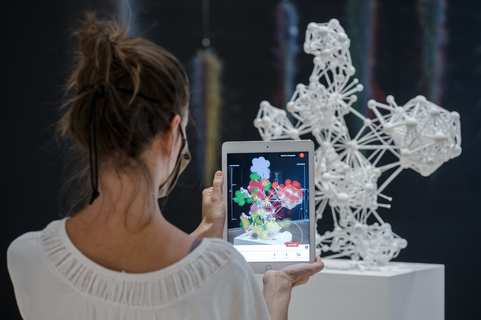 Eine Person steht vor einer weißen 3D-Netzwerk-Skulptur und hält ein Tablet hoch, auf dem mittels Augmented Reality die Skulptur näher untersucht werden kann.